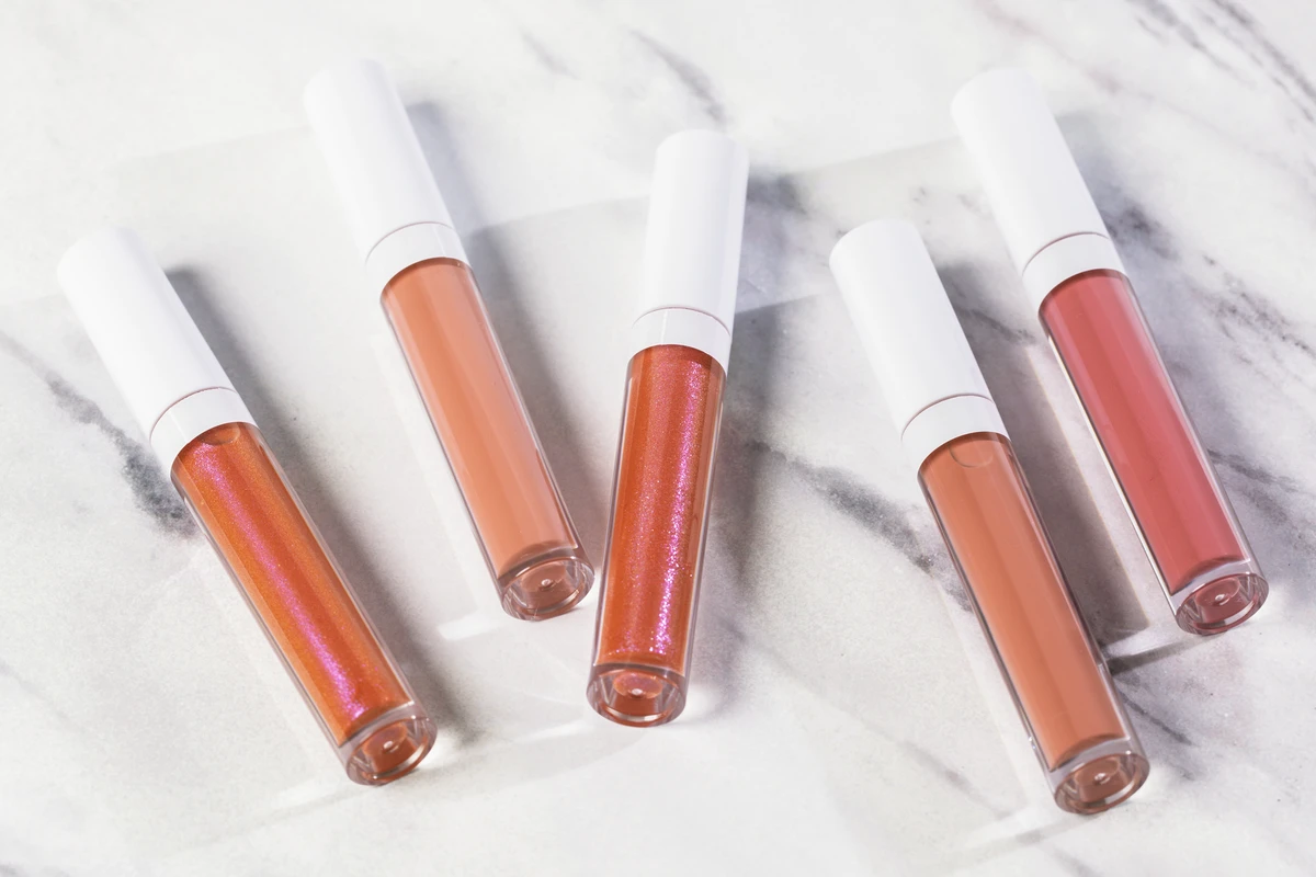 Close-up of 5 liquid blushes in clear bottles with white caps, featuring shades like clear, peach, and nude, set against a white marble-patterned background