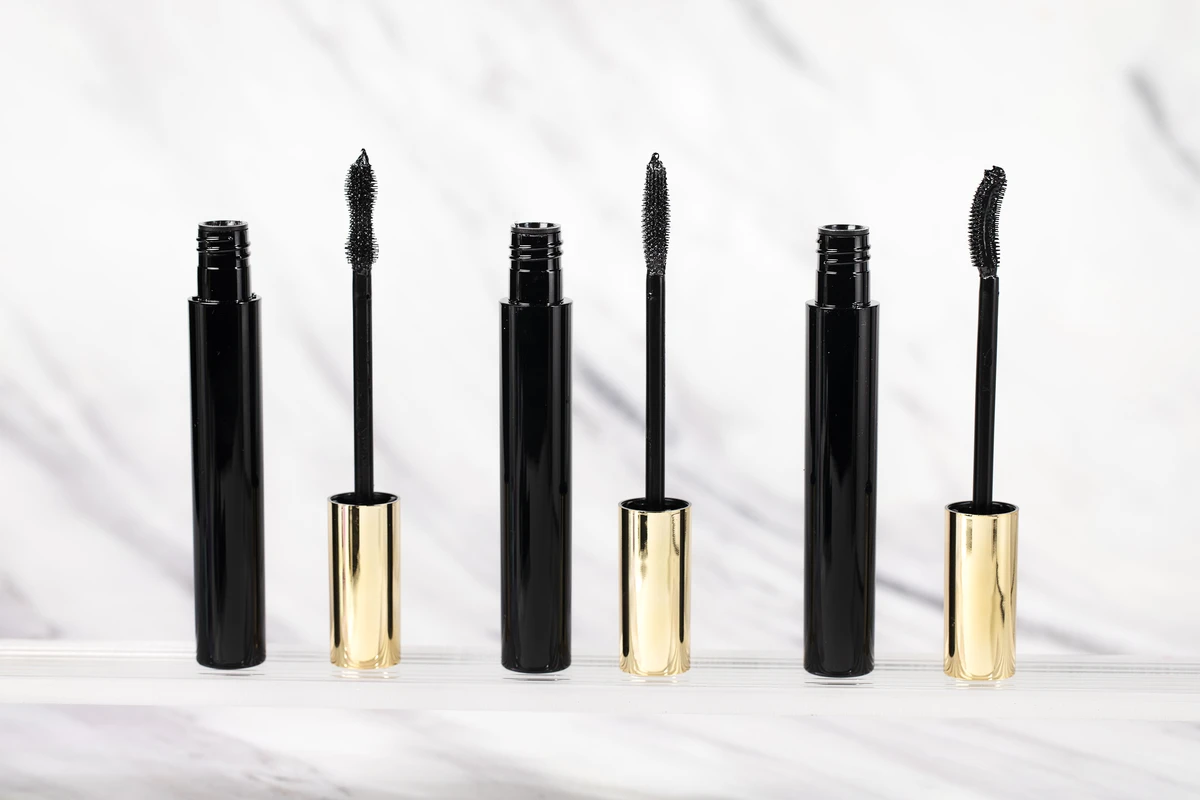 Close-up of 3 private label mascaras with black tubes and gold caps, each with a different brush shape for volume, curl, or length, set against a white marble-patterned background