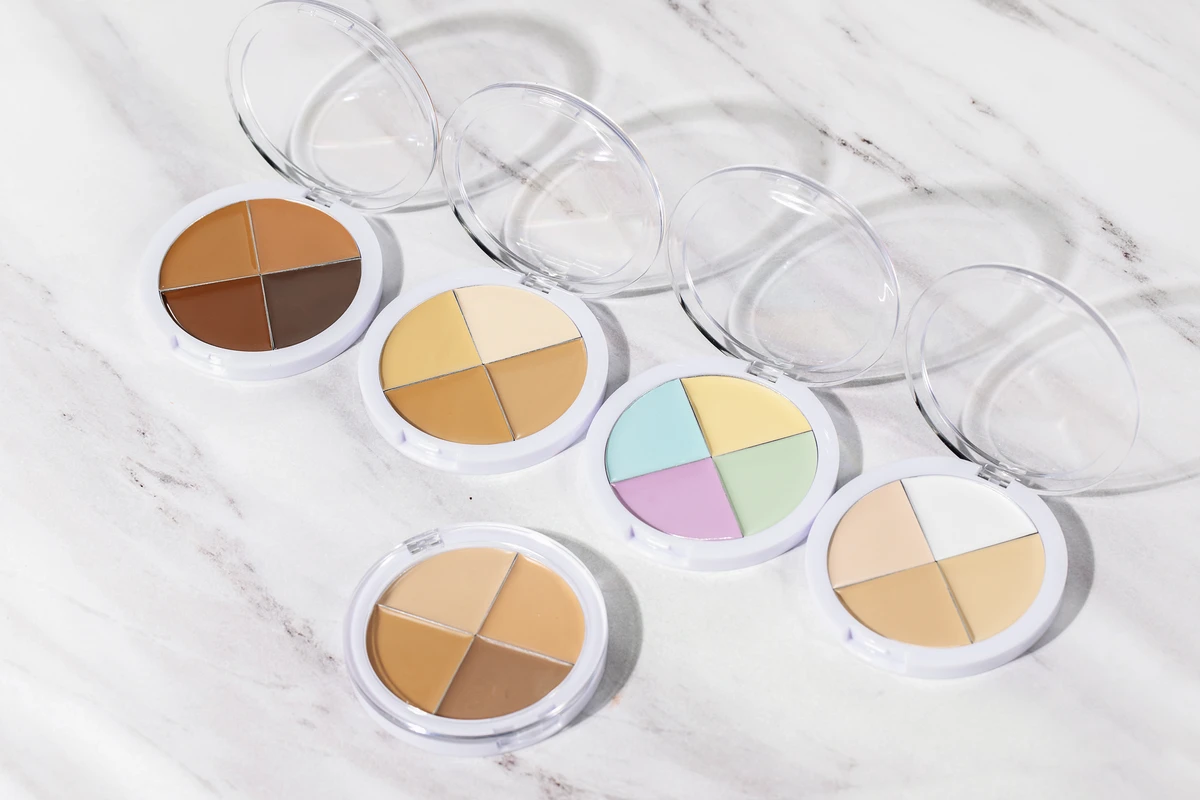Close-up of 5 four-color concealer palettes with clear jars, featuring shades like brown, clear, nude, skin tone, green, and light purple, set against a white marble-patterned background