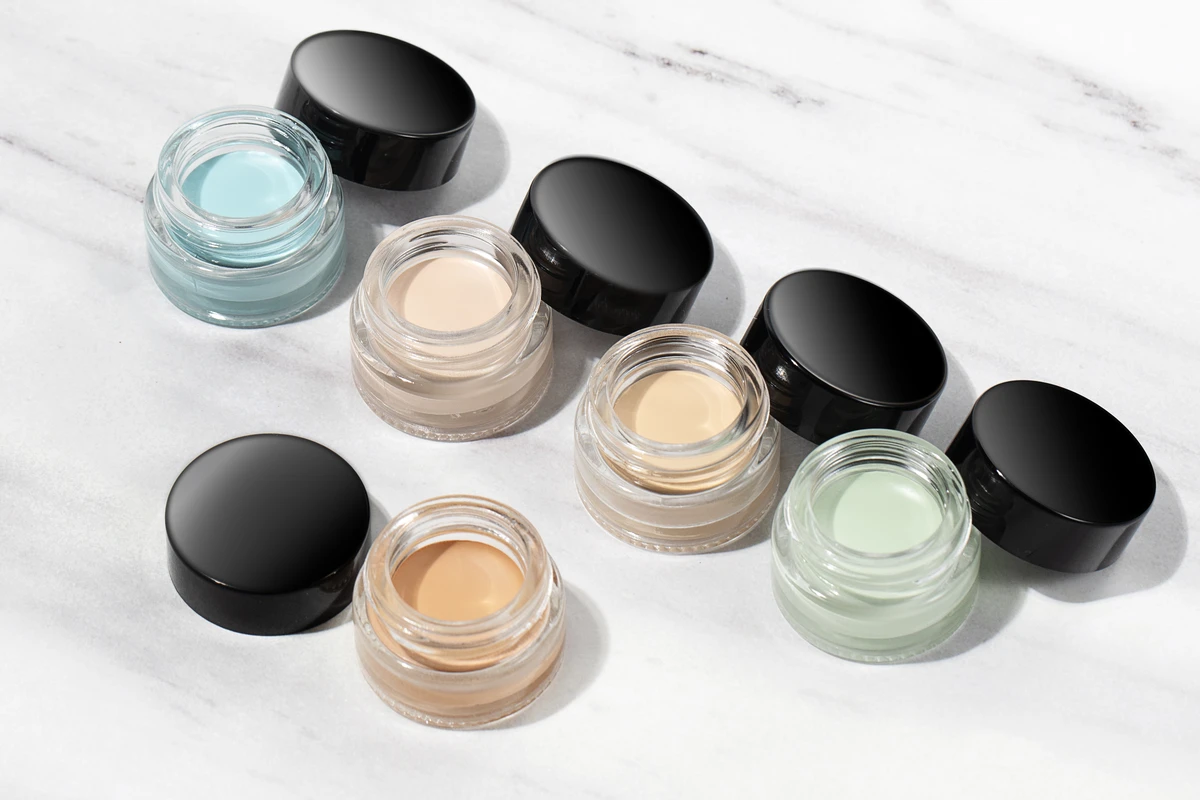 Close-up of 5 concealer pots with clear jars and black lids, featuring shades like nude, skin tone, and green, against a white marble-patterned background