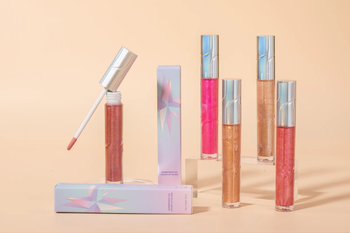 Five Ice Crush Lip Glosses in different shades (pink, magenta, light brown, etc.) with their colorful packaging boxes standing on acrylic columns. Light yellow background.