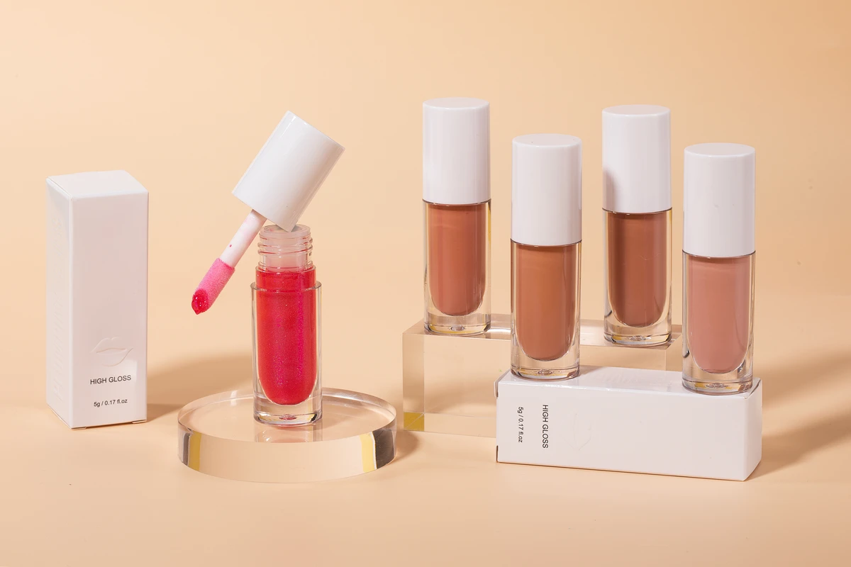 Five High Shine Lip Glosses in various shades (light pink, nude, etc.) with their white packaging boxes standing on acrylic columns. Light yellow background.