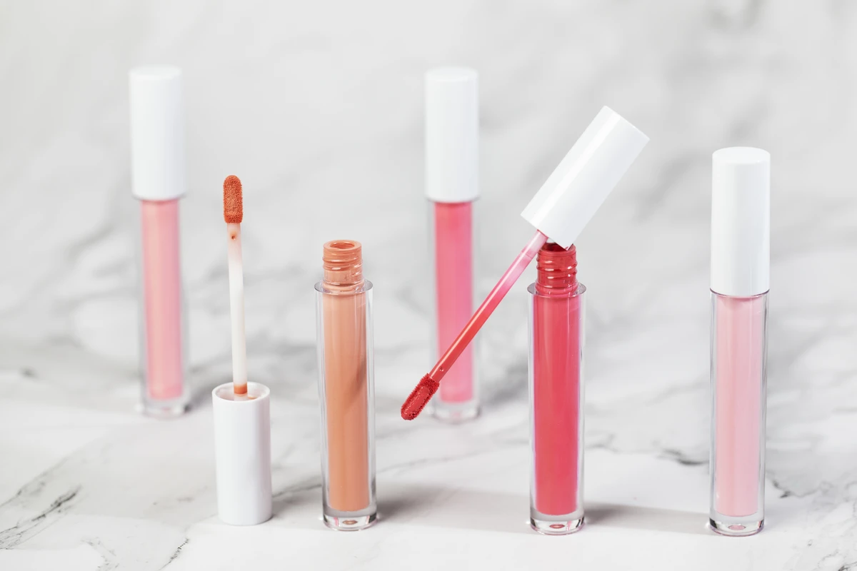 Close-up of private label lip gloss in peach and nude tones with round white tubes, glossy finish, and hydrating formula, set against a marble-patterned background.